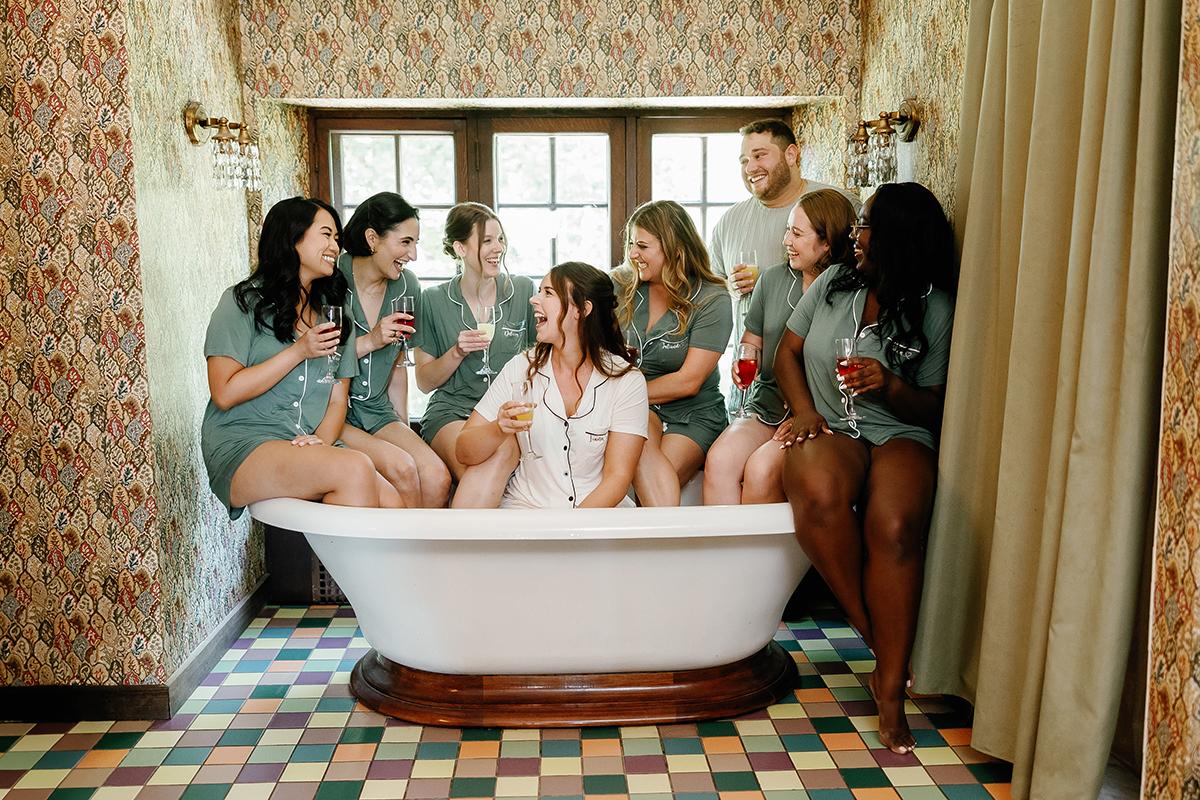Group of people laughing and sitting on a bathtub, holding drinks.