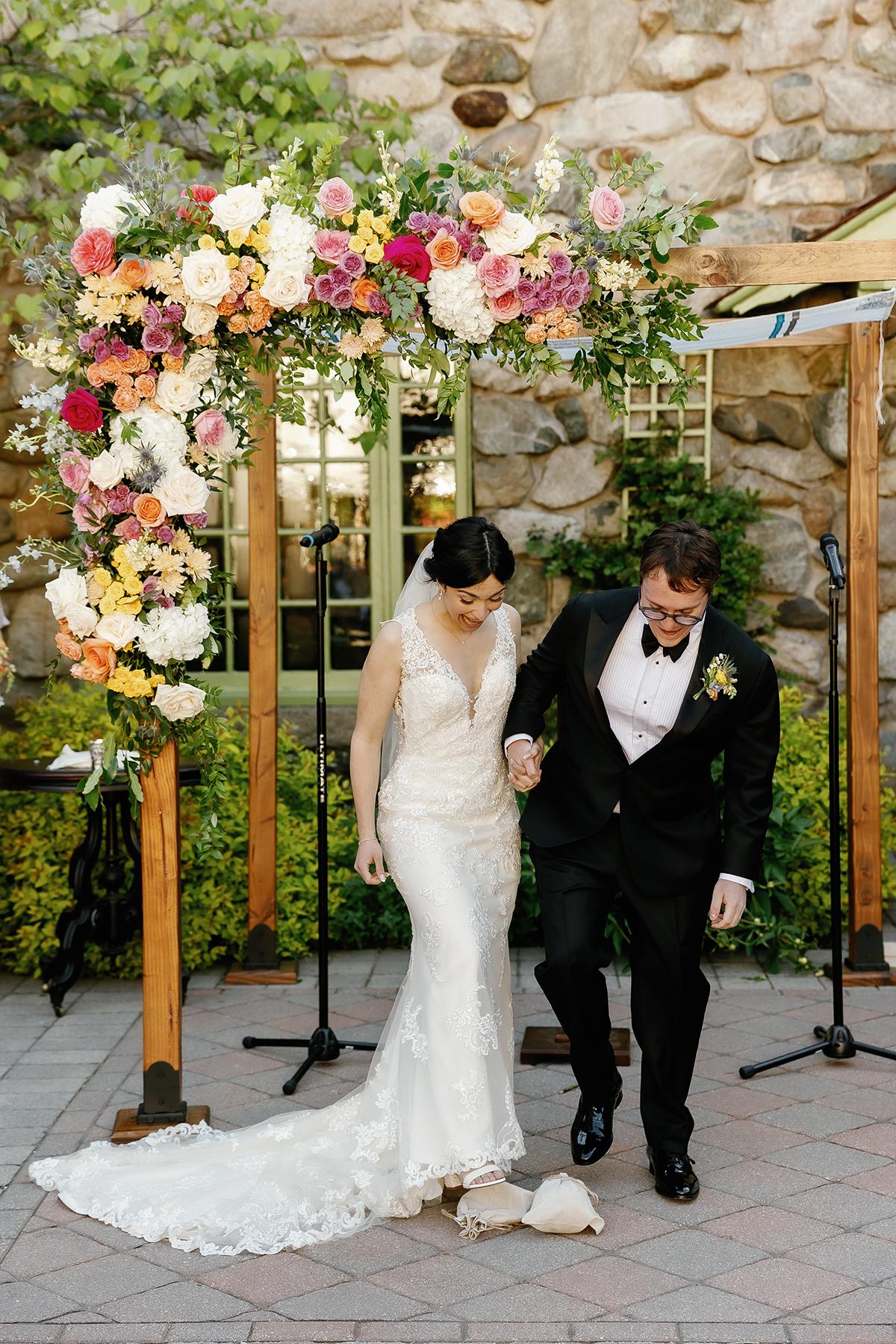 Bride and groom under floral arch, stepping forward together.