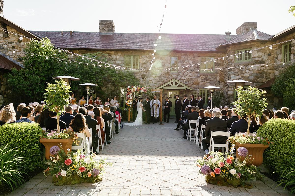 Wedding ceremony outdoors with guests seated, sunlight filtering through string lights.