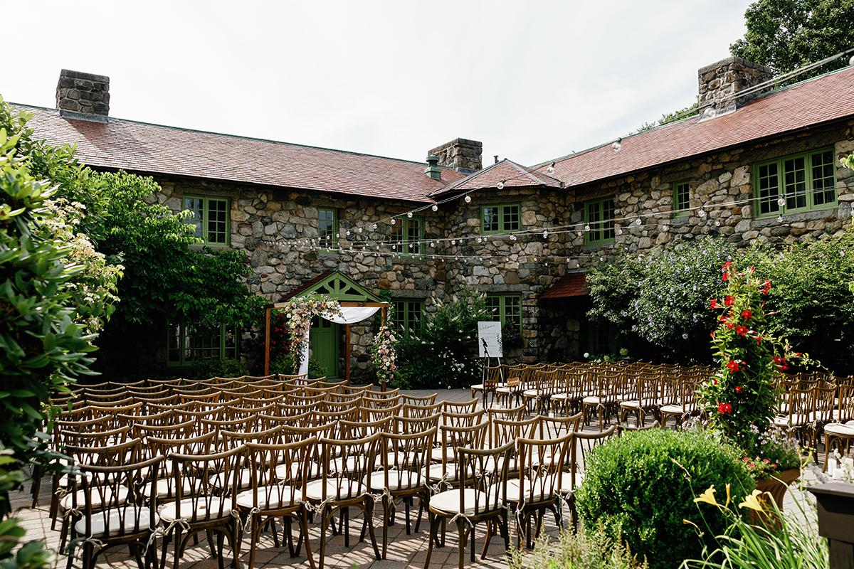 Courtyard set for a wedding with wooden chairs and stone building.