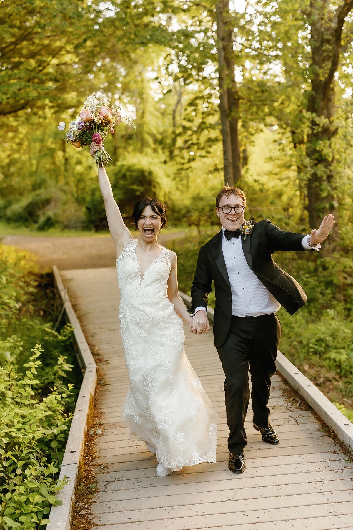Bride and groom joyfully walking on a wooden path in a forest setting.