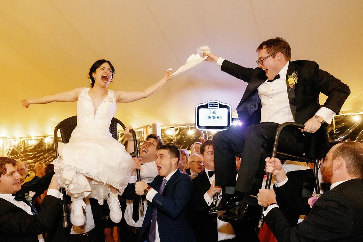 Bride and groom lifted in chairs, celebratory wedding scene.