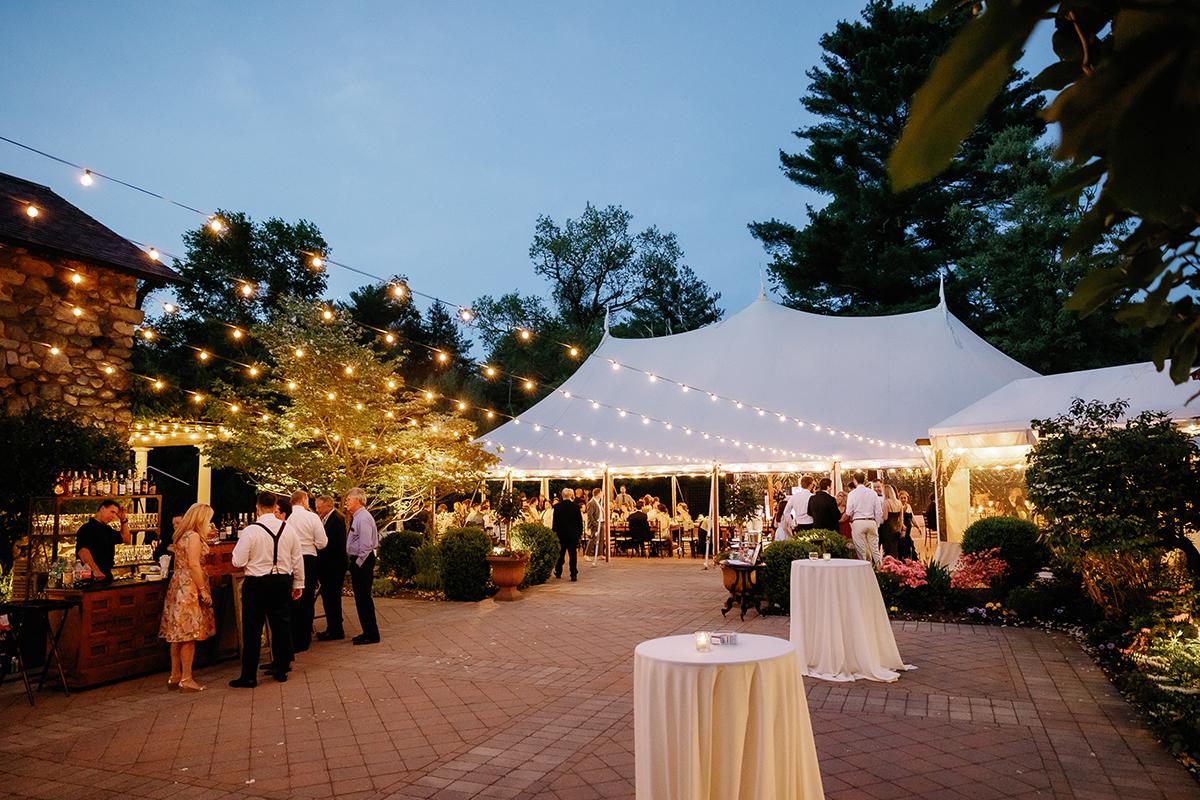 Outdoor wedding reception with string lights and guests mingling under a tent at dusk.