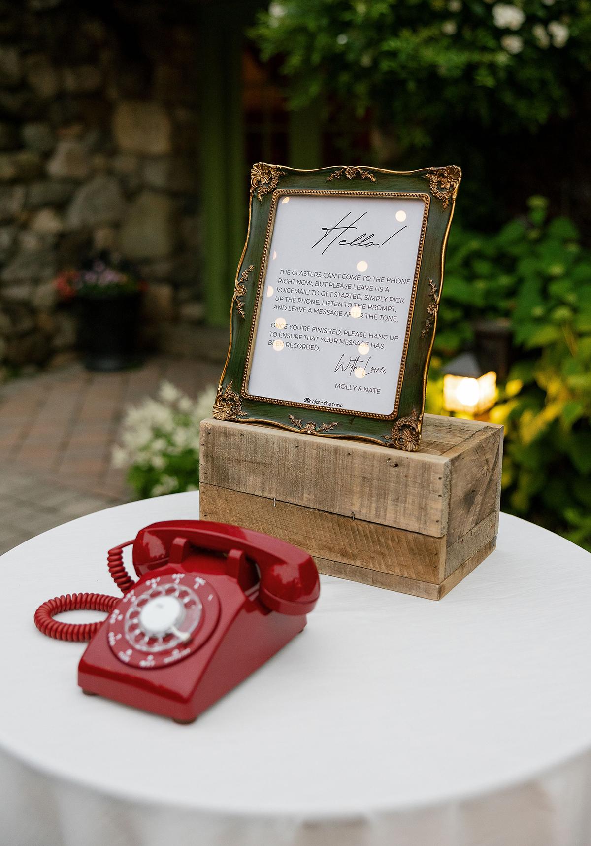 Red rotary phone on a table with a framed note in a garden setting.