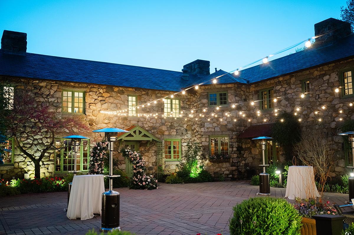 Courtyard with string lights, stone walls, and patio heaters at dusk.