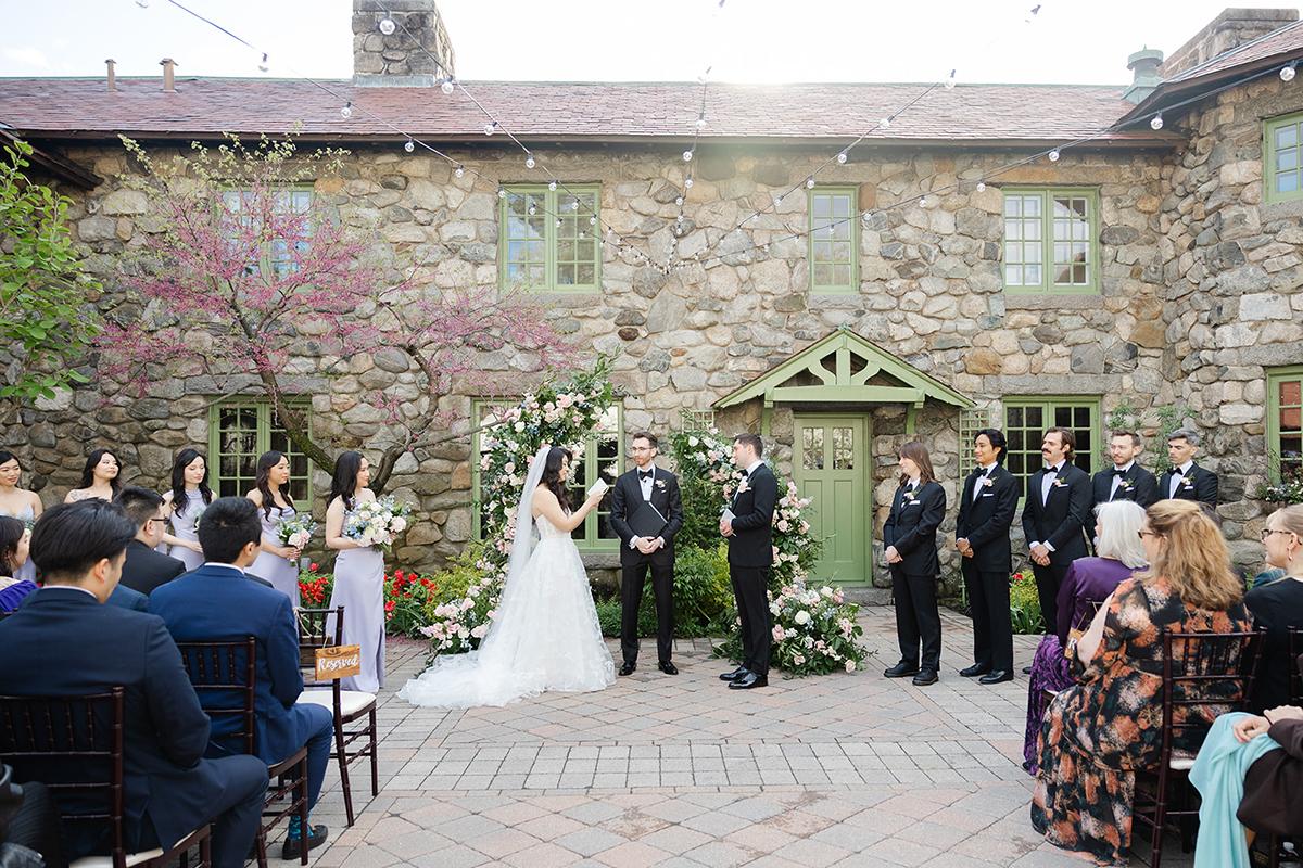 Bride and groom exchange vows outdoors, surrounded by wedding party and guests.