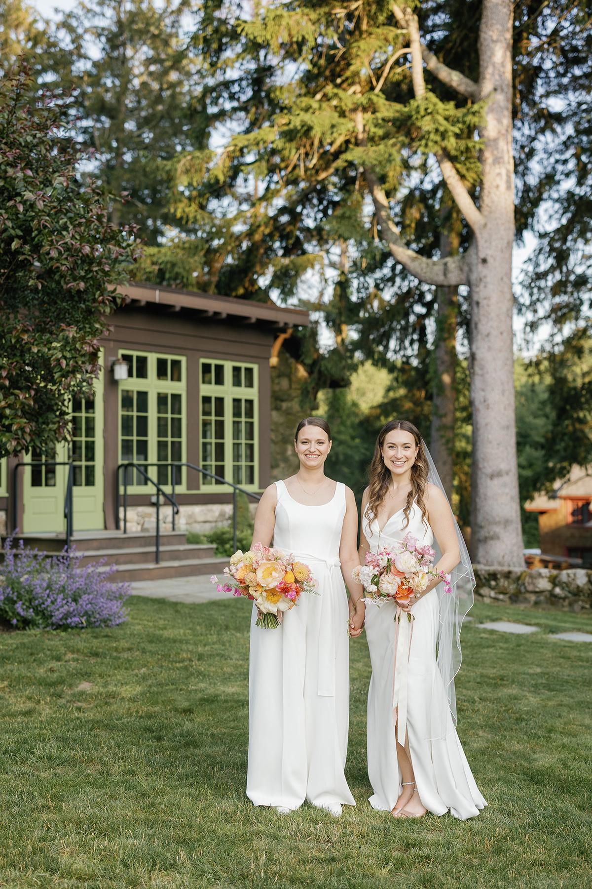 Two brides in white dresses holding bouquets, standing in a garden setting.