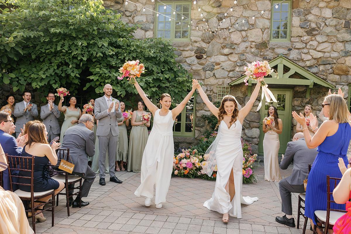 Two brides in white dresses celebrating with raised arms, surrounded by guests.