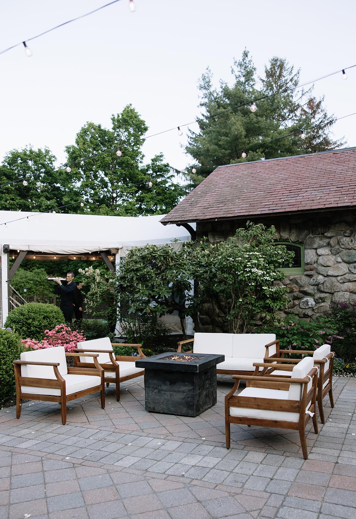 Patio with chairs, fire pit, and stone building surrounded by trees.