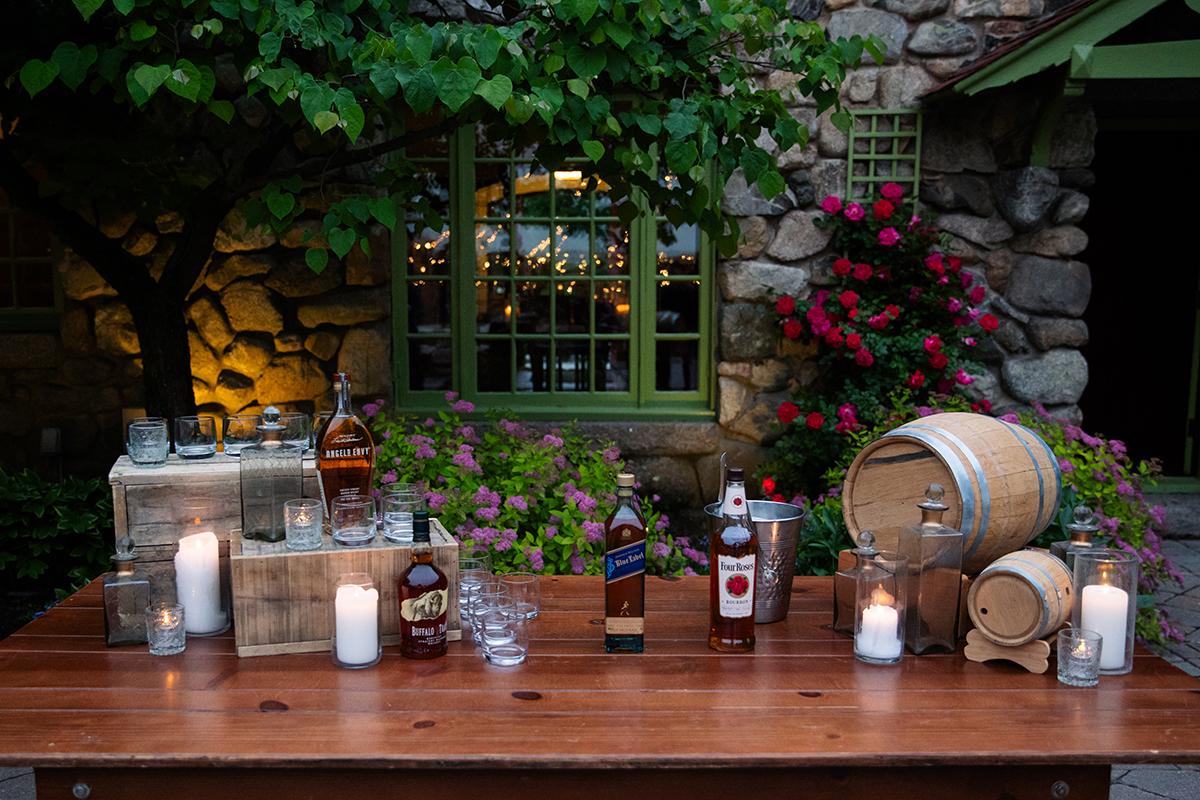 Outdoor bar setup with bottles, barrels, and candles on a wooden table. Stone wall, greenery, flowers.