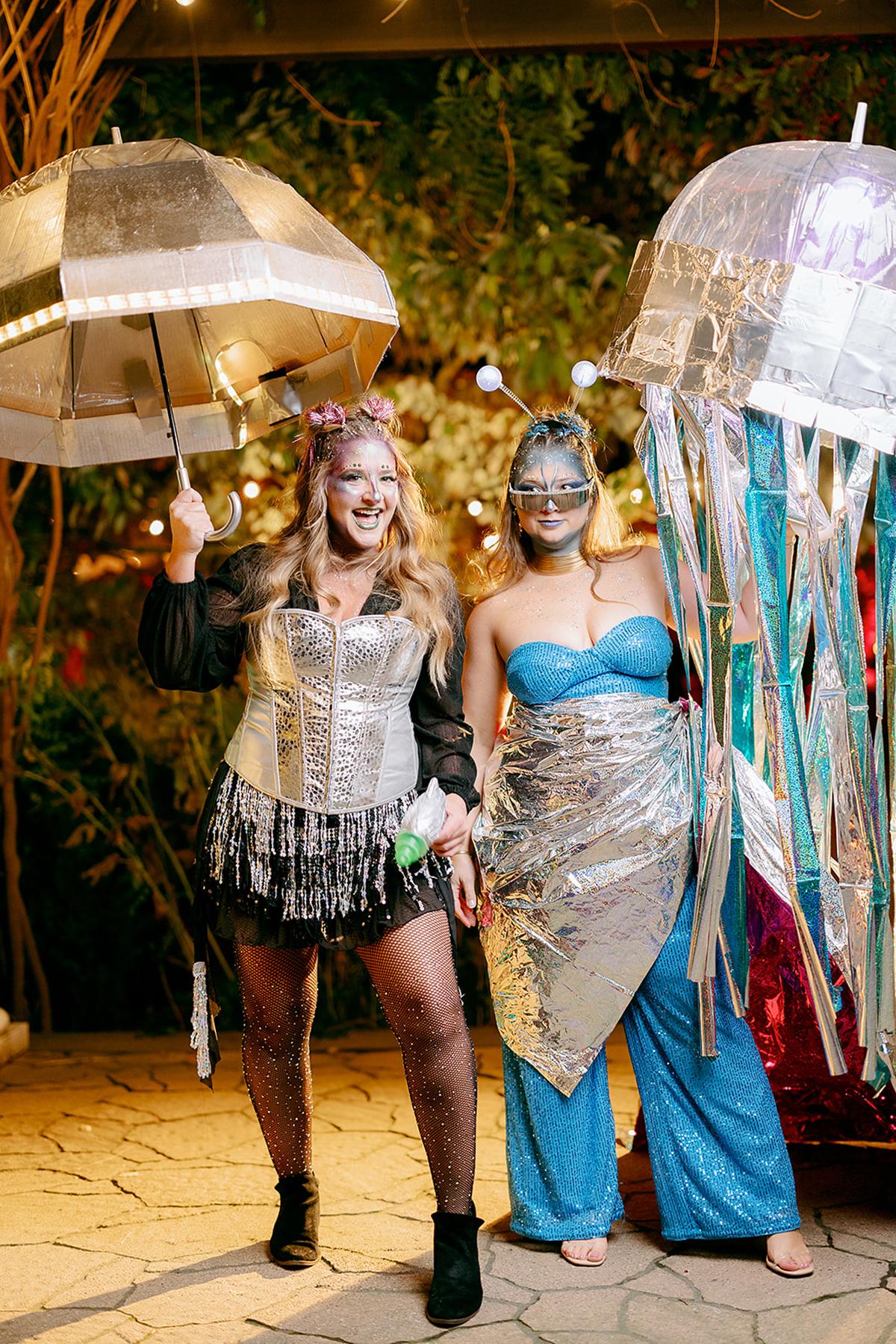 Two people in creative costumes under dim lights outdoors, one with a metallic umbrella.