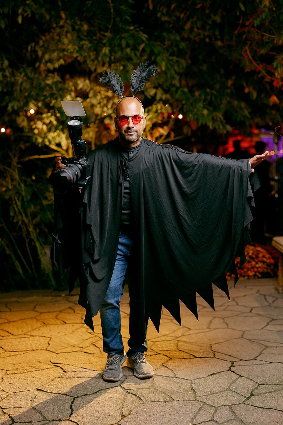 Man in black costume with bat-like wings and red sunglasses, standing outdoors at night.