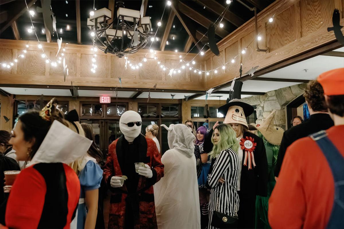Party guests in Halloween costumes gather under string lights indoors.