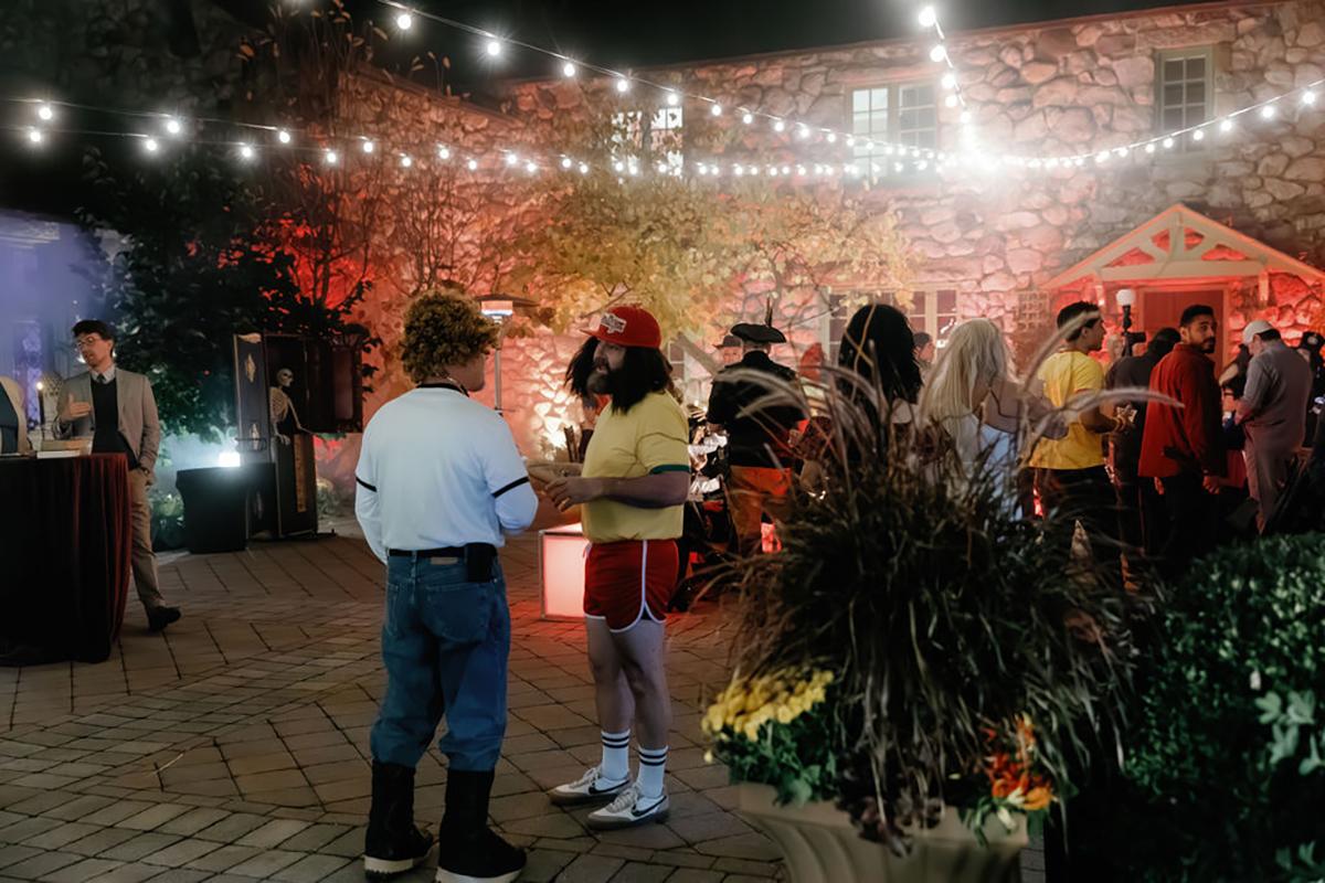 Crowd at a nighttime outdoor party with string lights and autumn decorations.