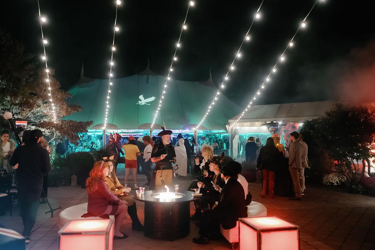 Outdoor night party with string lights, people socializing and seated around round tables.