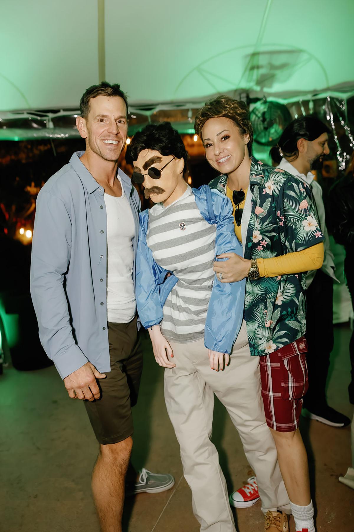 Three people in festive attire, one holding a mannequin, smiling under green party lights.