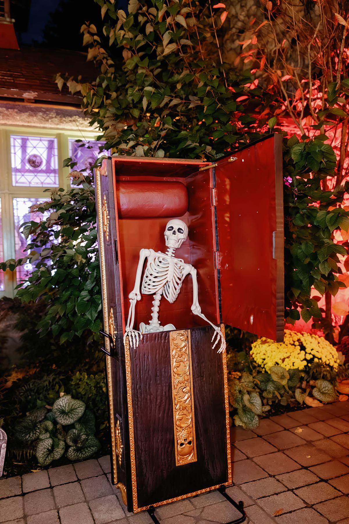 Skeleton in an upright coffin with colorful lights and plants in the background.