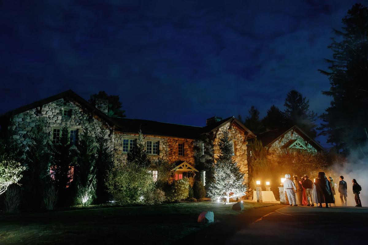 Large stone house at night, warmly lit with people gathered outside.
