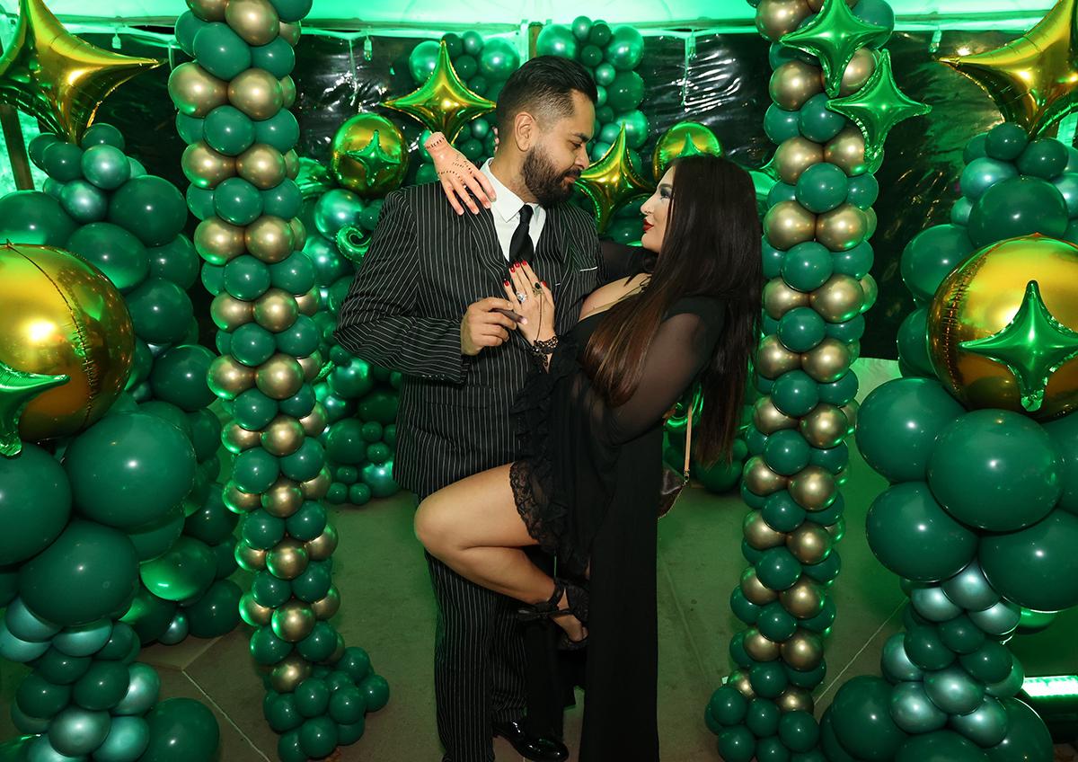 Couple posing in a balloon arch decorated with green and gold balloons.