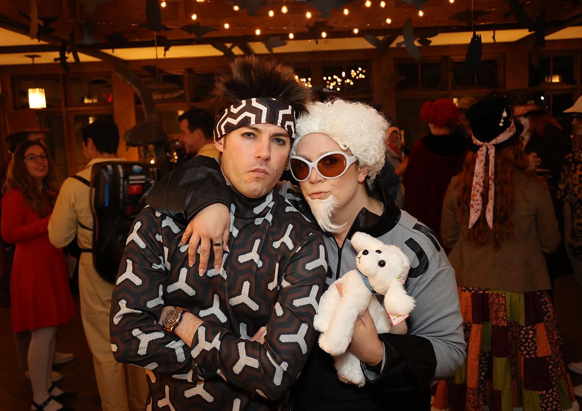 Two people in costume at a party, one holding a plush toy, under string lights.