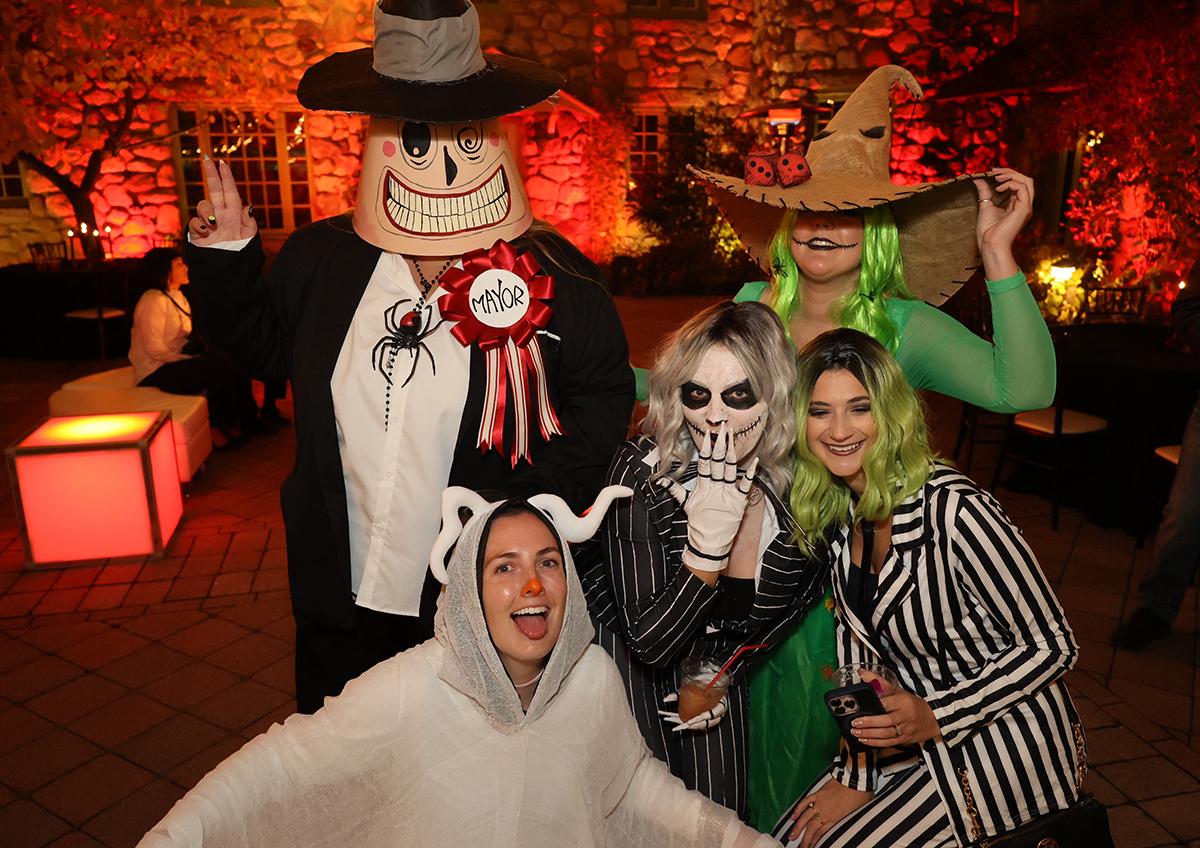 Five people in Halloween costumes, smiling in a warmly lit outdoor setting.