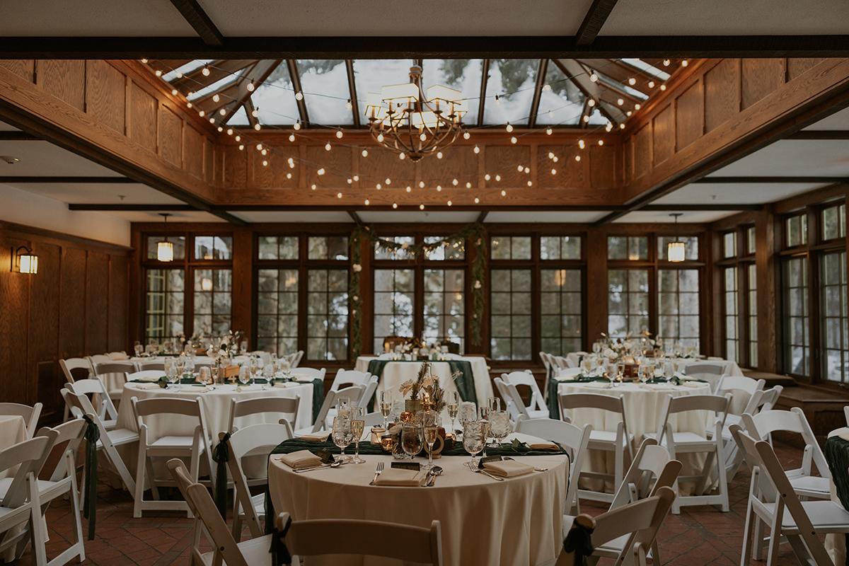 Elegant banquet room with round tables, white chairs, and string lights overhead.