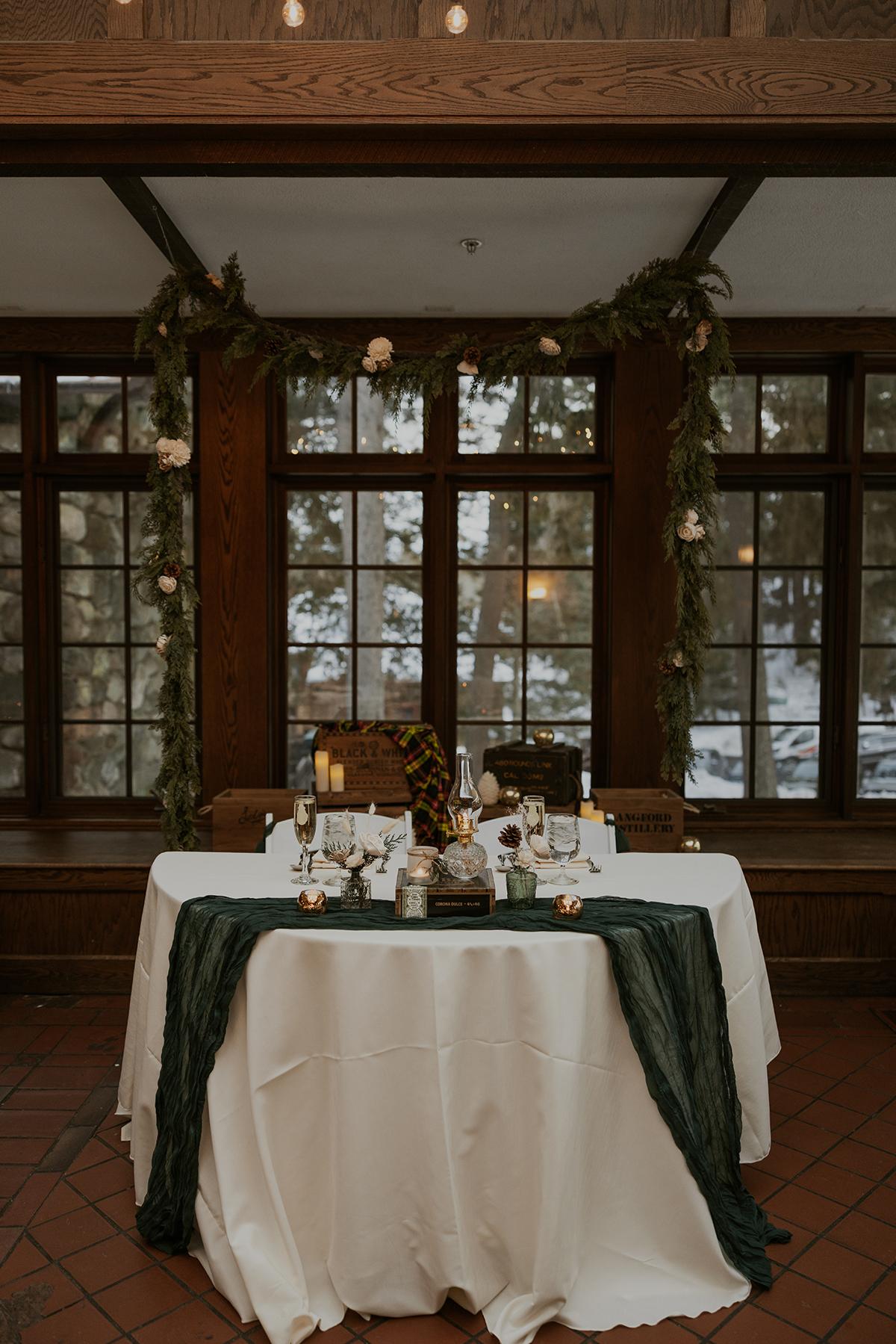 Elegant dining table set with candles and greenery, in front of large windows.