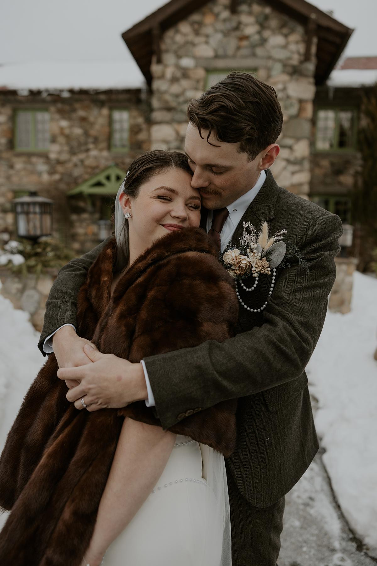 Couple hugging outdoors in winter attire, smiling in front of a stone house.