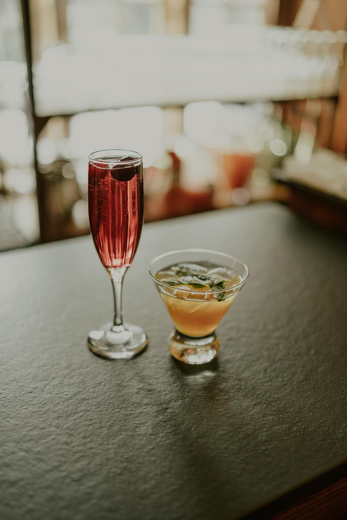 Two cocktails on a bar counter, one red, one orange with mint leaves.