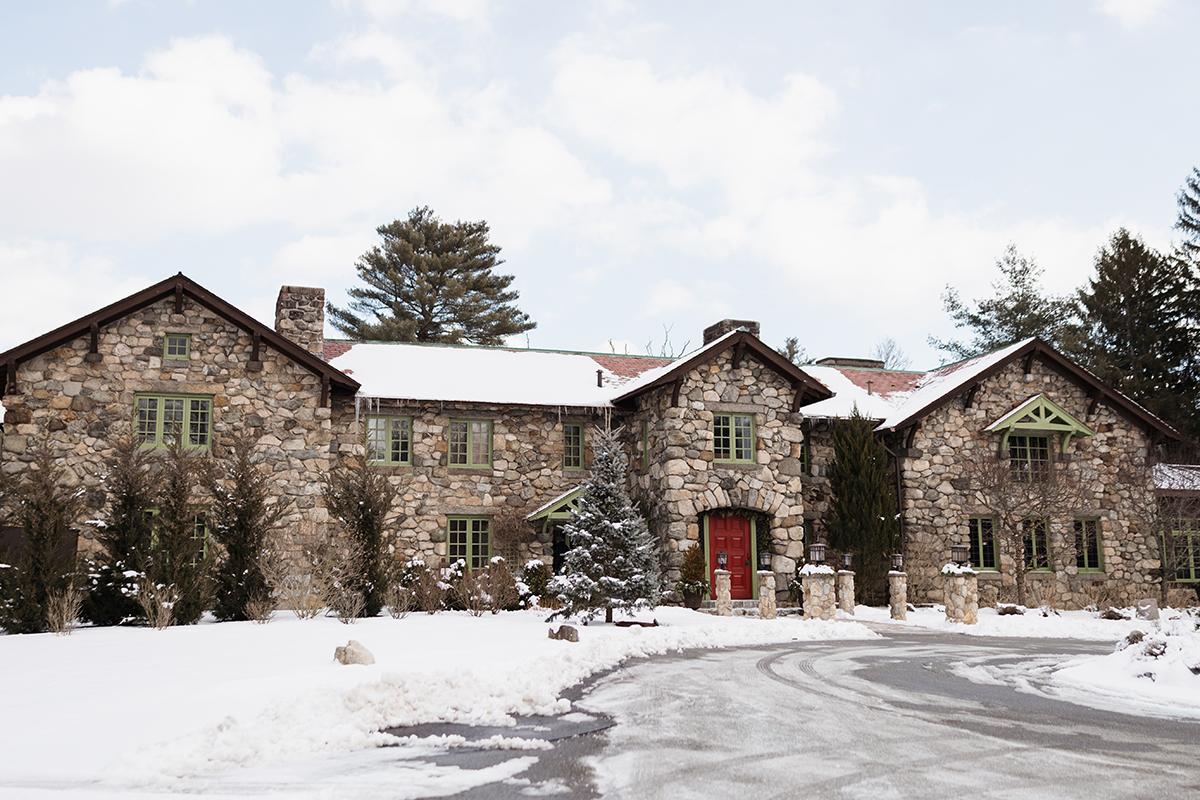 Stone mansion with green windows, red door, surrounded by snow-covered ground and trees.