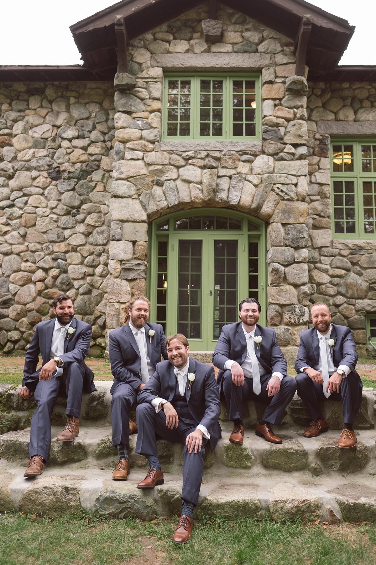 Men in suits sitting on stone steps, posing in front of a rustic stone building.