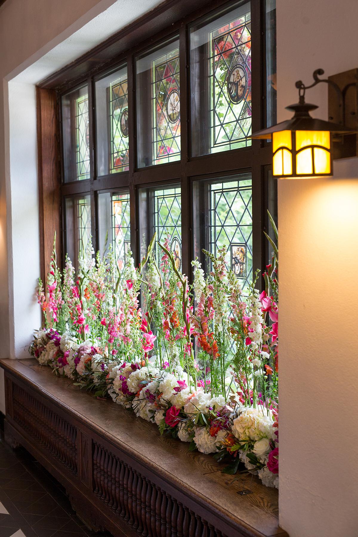 Stained glass windows with colorful flowers on a wooden ledge, softly lit by a wall lamp.