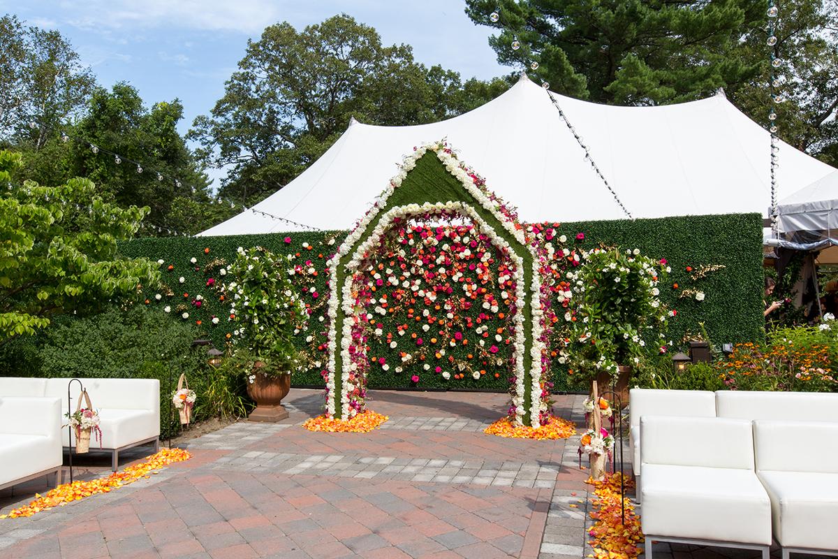 Wedding arch with colorful flowers under a white tent, surrounded by greenery.