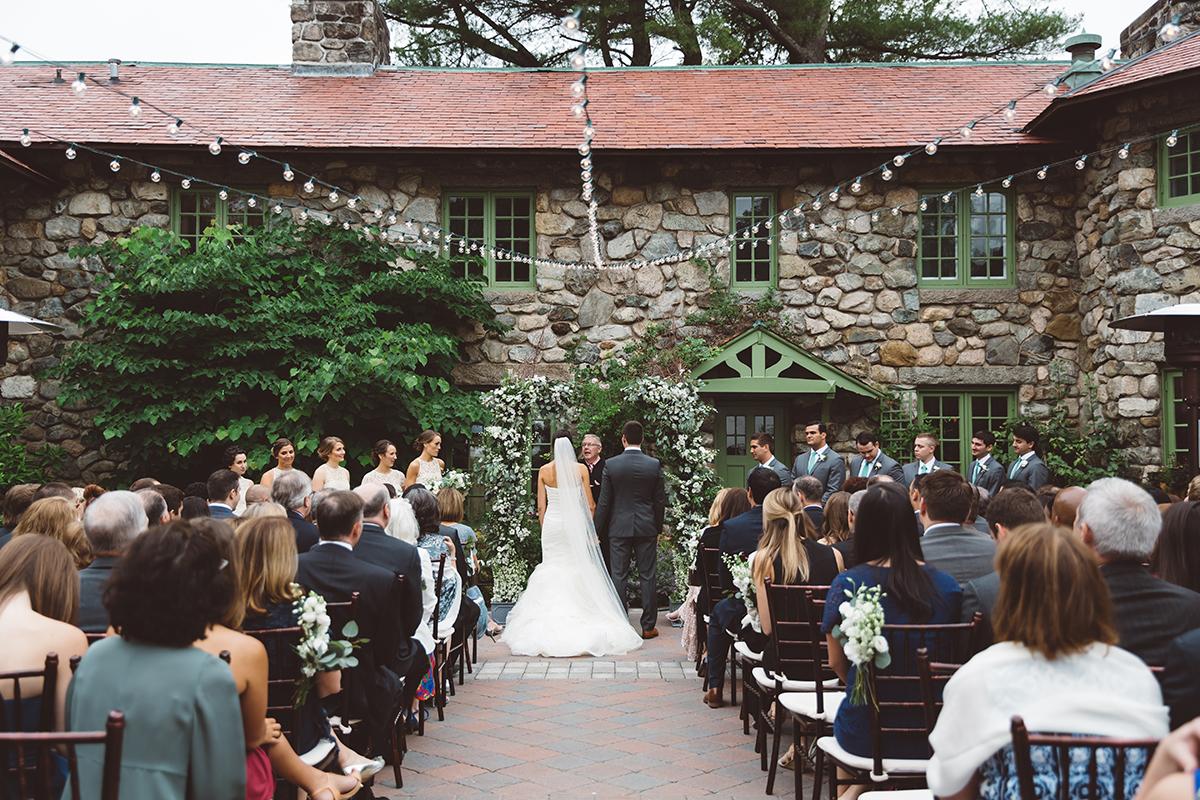 Outdoor wedding ceremony at a stone building, with bride and groom facing officiant.