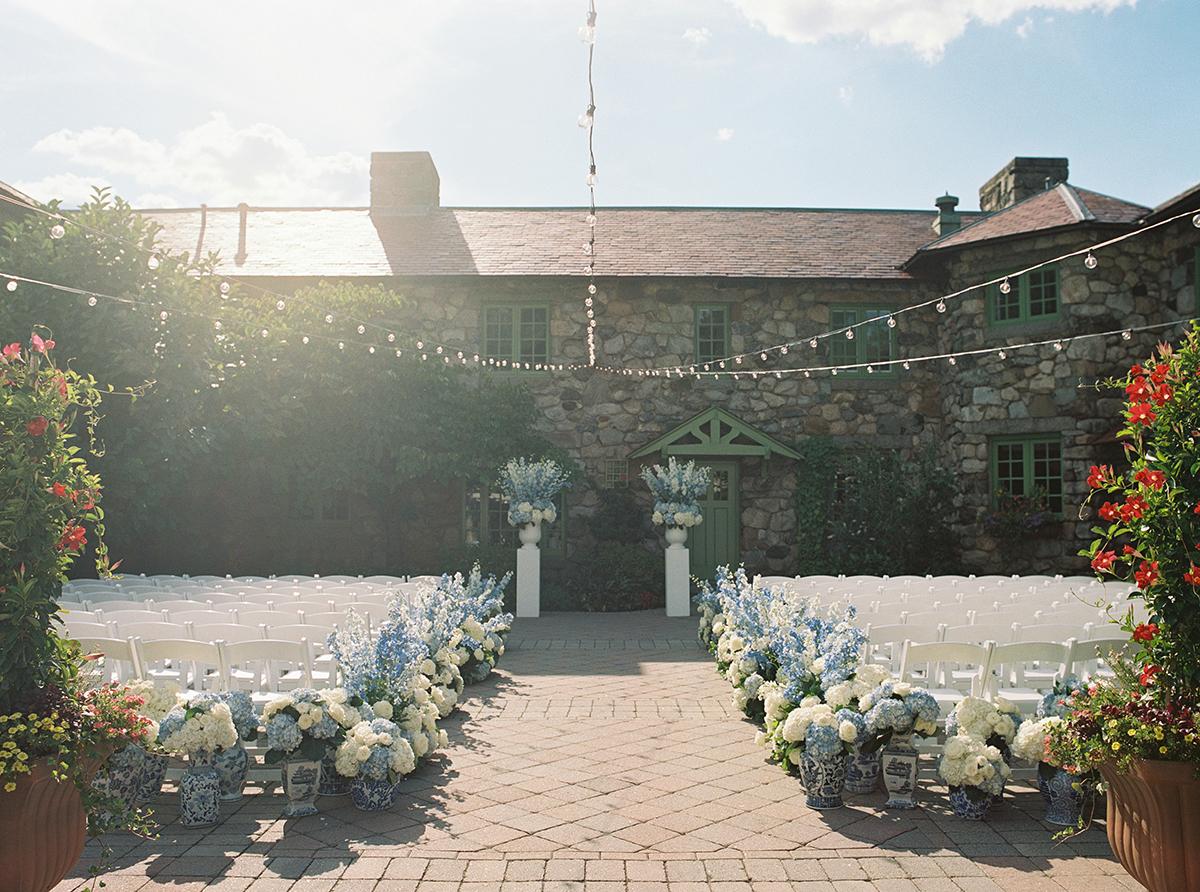 Courtyard wedding setup with chairs, flowers, and a rustic stone building in sunlight.