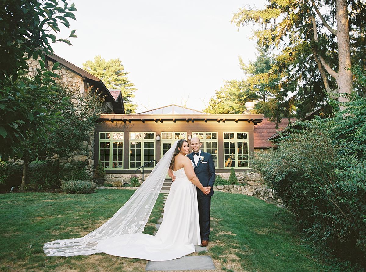 Bride and groom in garden, surrounded by greenery and a building.