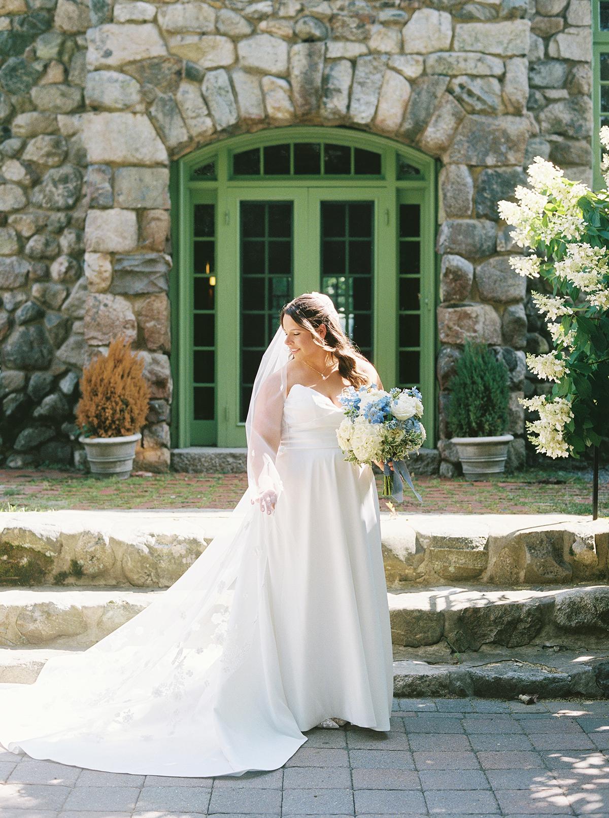 Bride in white dress holding bouquet, standing by stone wall and green doors.