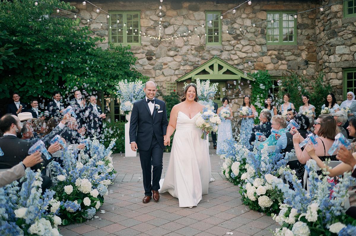 Bride and groom walk down aisle, surrounded by guests and blue flowers. Stone building backdrop.