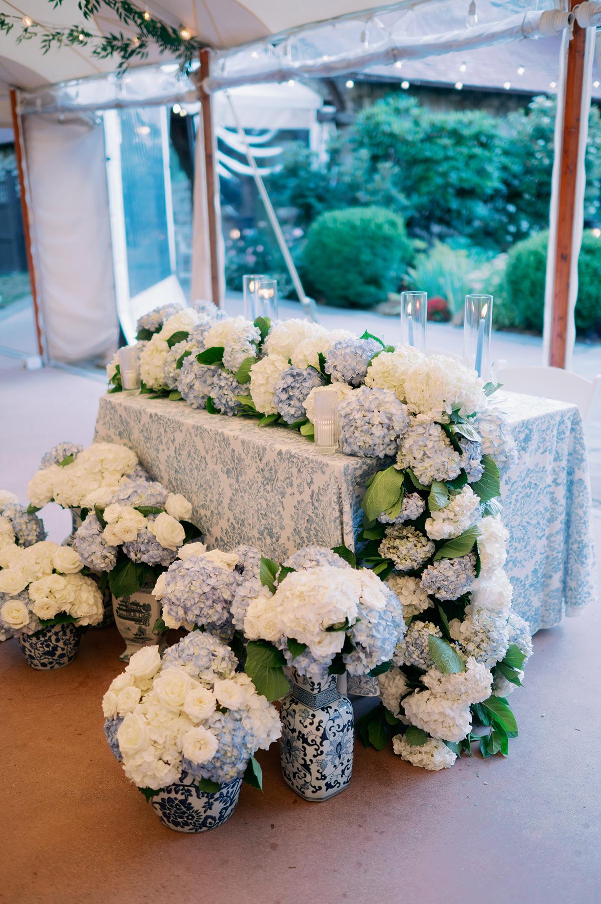 Flower-adorned table with white and blue hydrangeas under a tent.