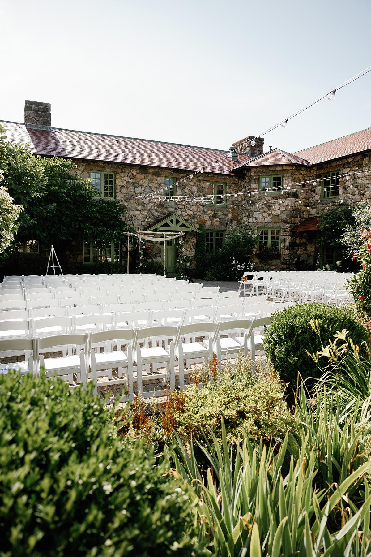 Outdoor wedding setup with white chairs facing a rustic stone building.