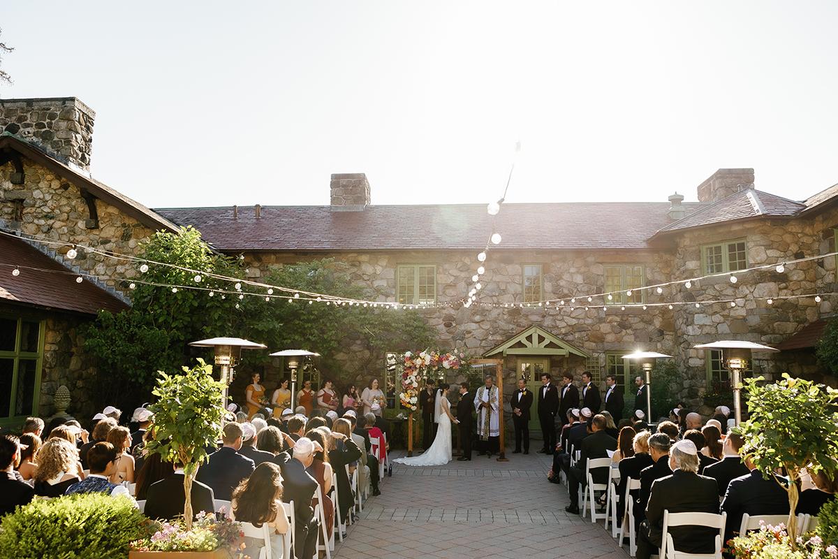 Outdoor wedding ceremony with guests seated, sunny day, string lights overhead.