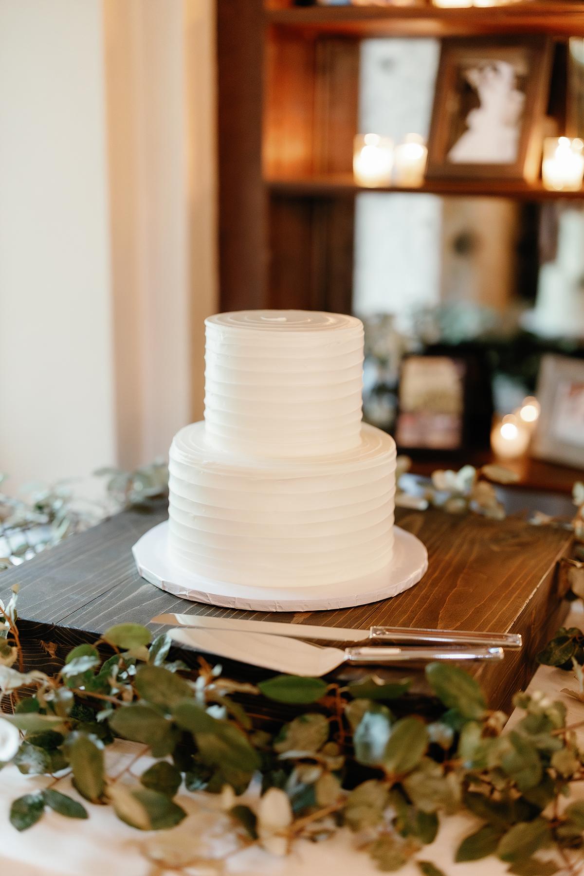 Two-tier white cake on a wooden stand, surrounded by greenery.
