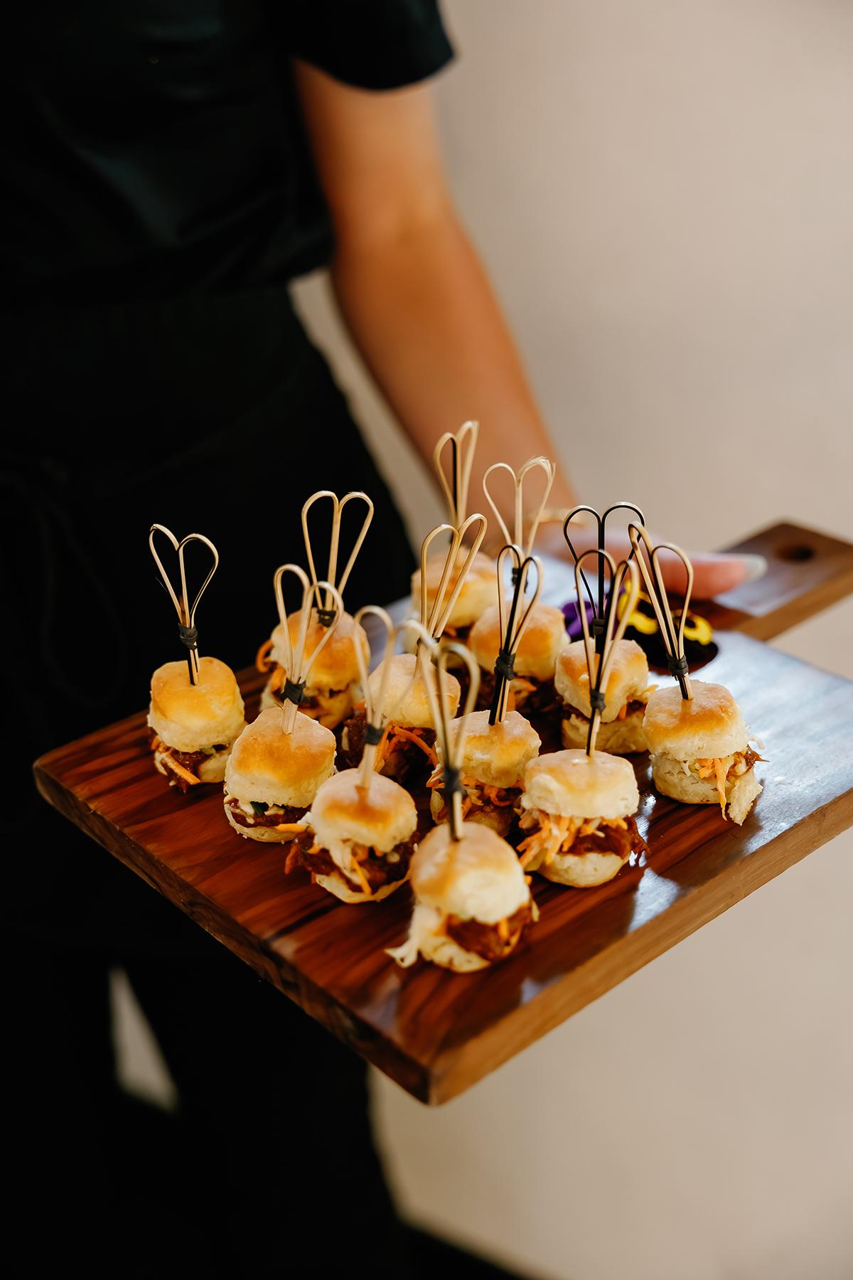 Mini sliders on a wooden tray held by a person in black attire.