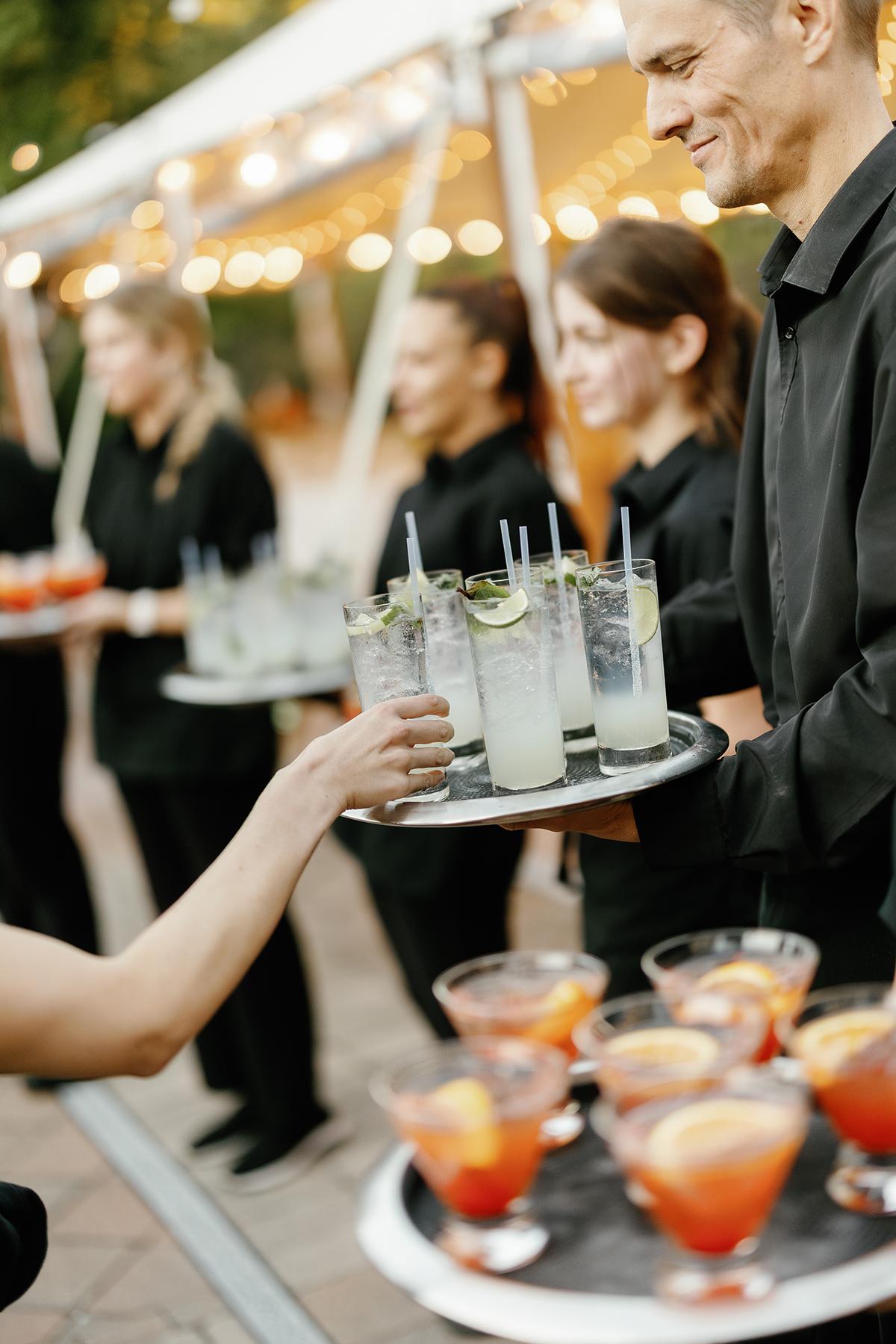 Servers in black uniforms handing out drinks on trays at an outdoor event.