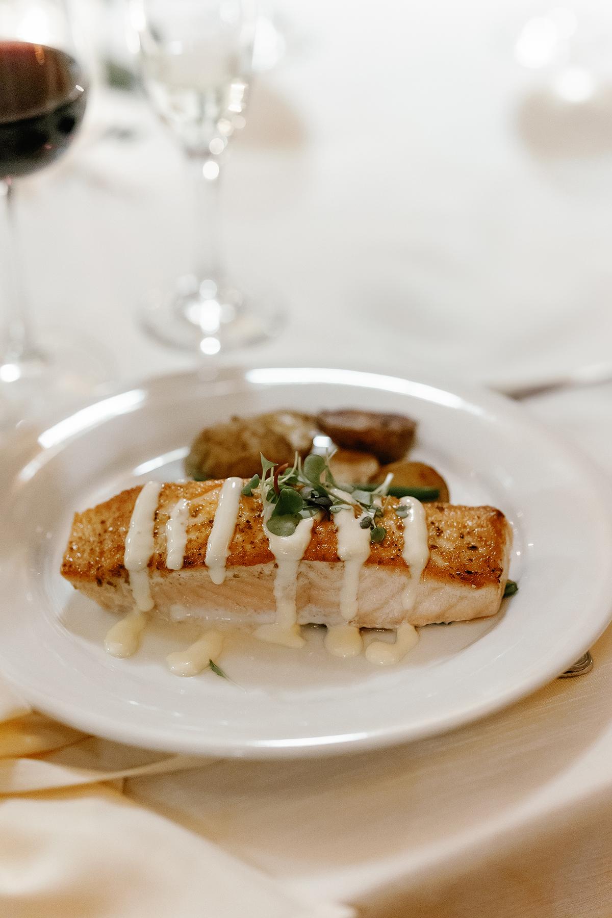 Grilled salmon with sauce and herbs on a white plate, with wine glasses in the background.