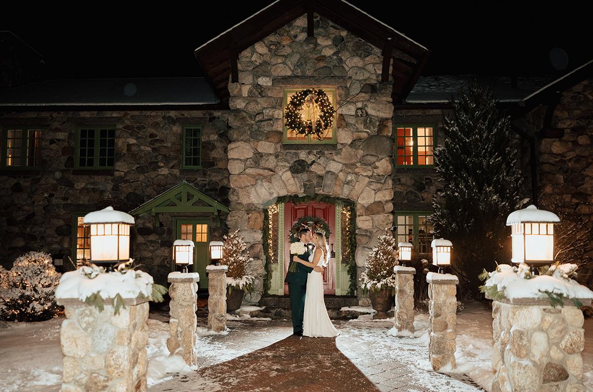 Bride and groom embrace outside a snowy, stone house at night.