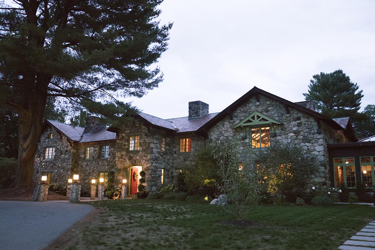 Stone house with warm lights, surrounded by trees at dusk.