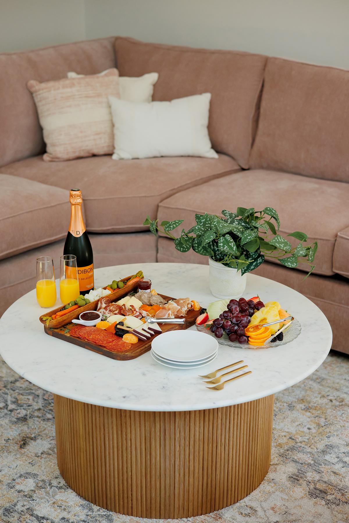 Champagne, fruit, and pastries on a white table in front of a pink sofa.