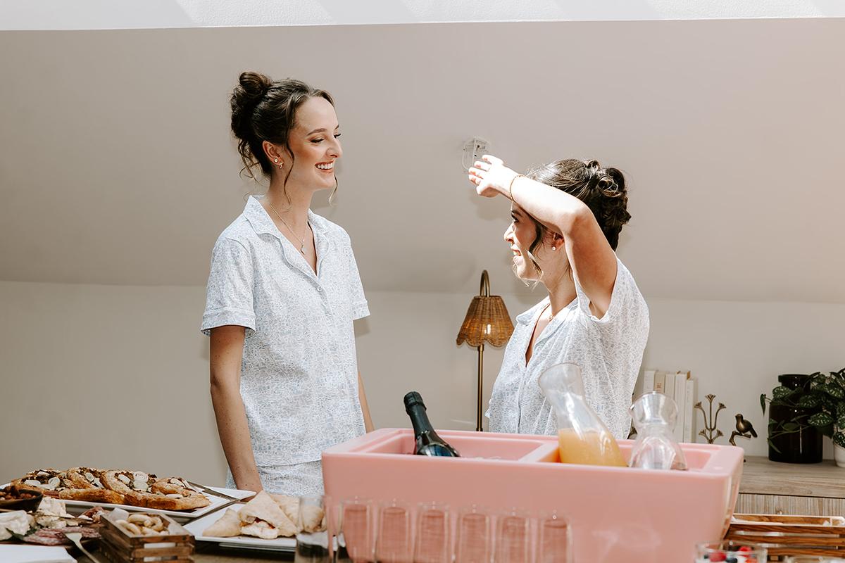 Two women in pajamas chatting by a kitchen counter with snacks and drinks.