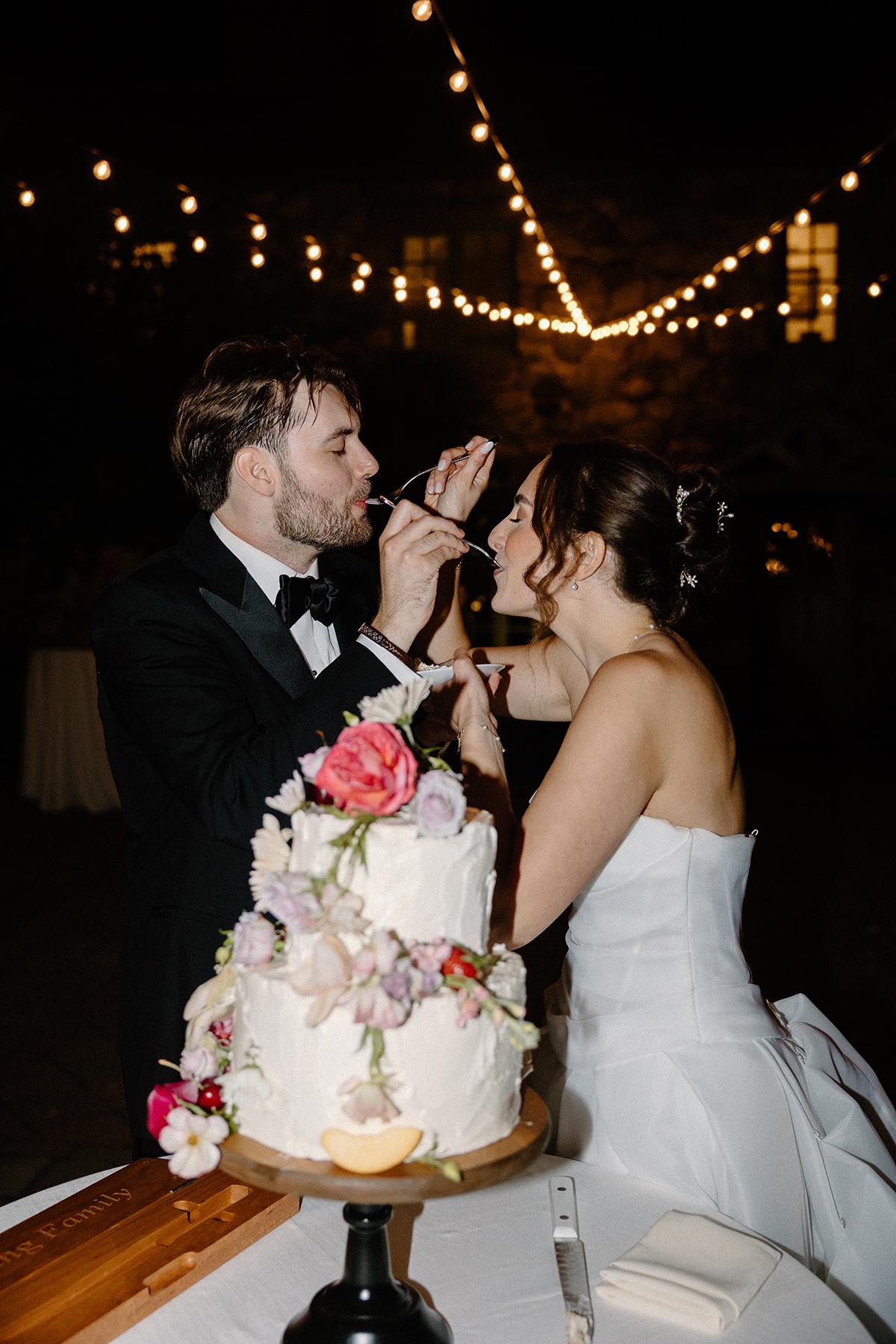 Newlyweds feeding each other cake under string lights at night.